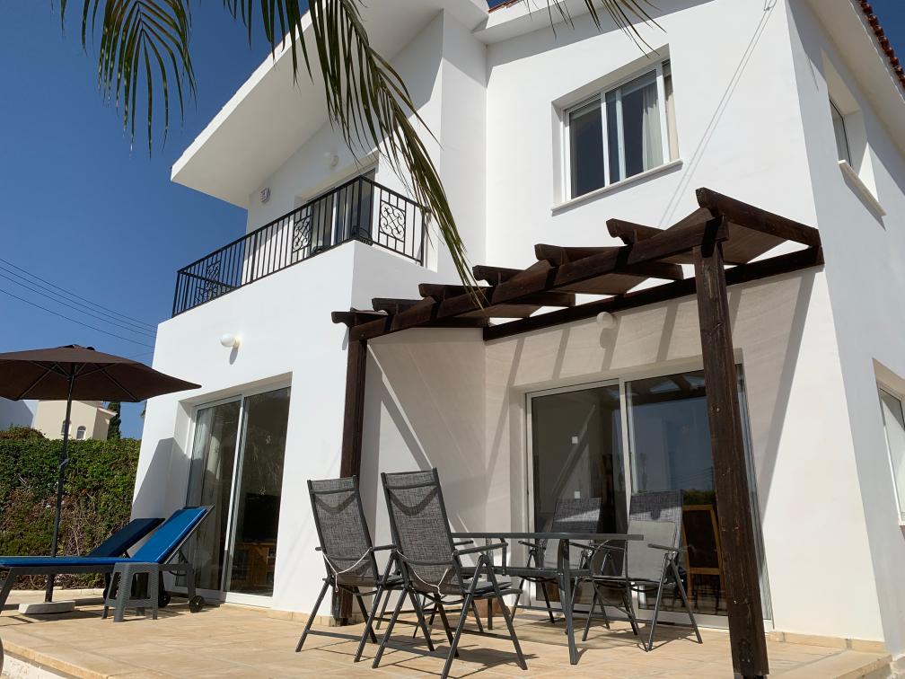 The white two-storey villa in sunshine, with a balcony above and a wooden pergola shading a terrace table and chairs.